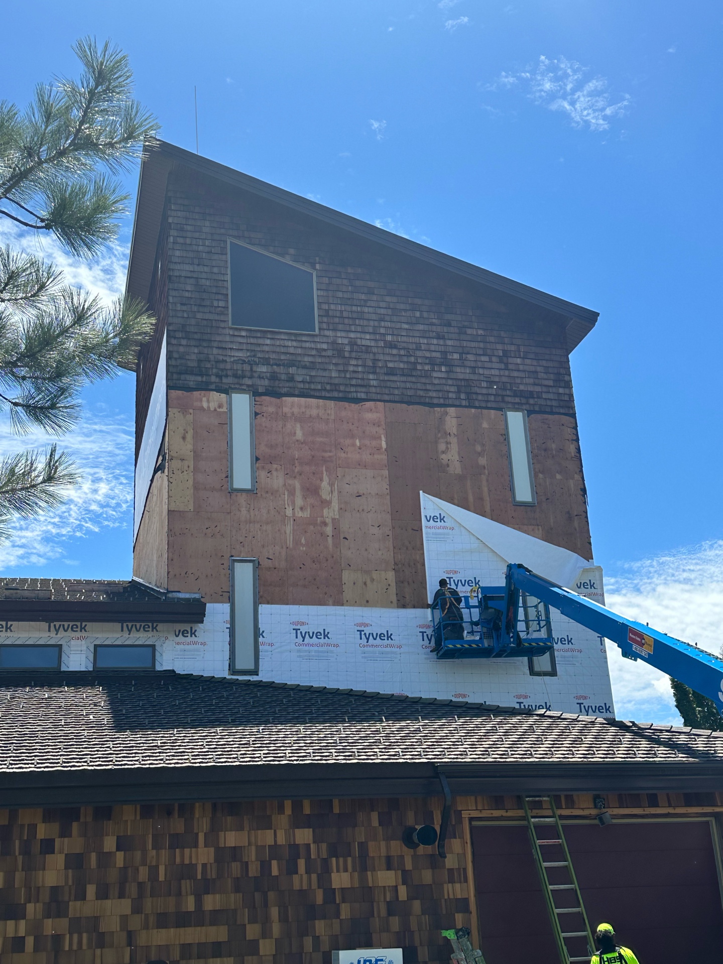 During photo of McCall Smoke Jumper Base tower — crew on boom lift installing new cedar shake siding on upper section with Tyvek visible below